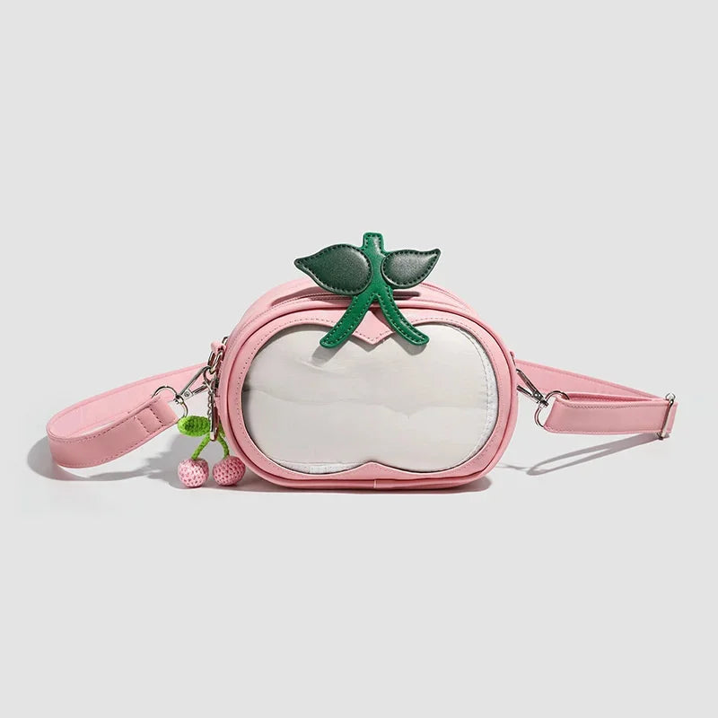 Apple-Shaped Crossbody Phone Purse – Lightweight Shoulder Bag A pink faux-leather crossbody bag designed to look like a peach, featuring a clear front window, a green leaf and stem accent on top, and two small dangling cherry charms on the side strap, set against a light gray background.