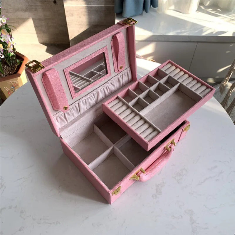 Leather Travel Jewelry Box with Mirror – Double-Layer Jewelry Organizer A pink hard-shell jewelry case shown open on a light marble-like surface, with interior compartments, a mirror, and a sliding tray visible. The case has gold-tone hardware and is photographed in a real indoor setting with sunlight and surrounding furniture visible.