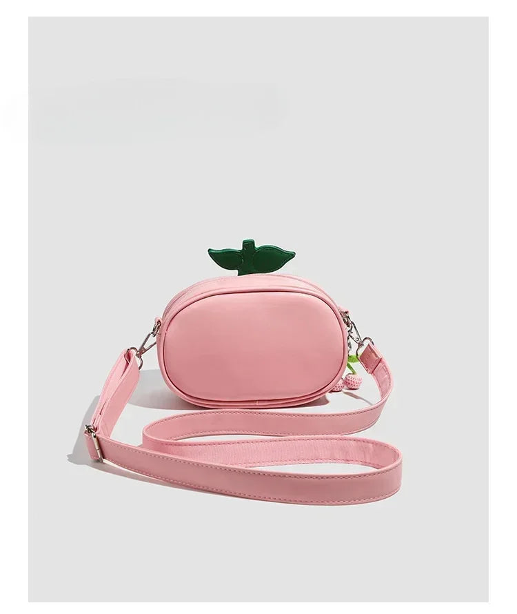 Apple-Shaped Crossbody Phone Purse – Lightweight Shoulder Bag A pink faux-leather crossbody bag designed to look like a small peach or fruit, featuring a green leaf on top and a matching pink shoulder strap, shown against a light gray background.