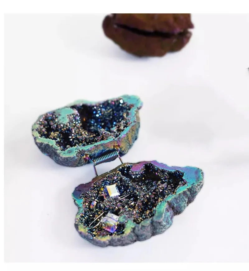 Luxury Portable Jewelry Box – Ring & Earring Travel Storage Case A pair of miniature jewelry display boxes shaped like open geode specimens, featuring iridescent turquoise and purple outer layers, dark glittery interiors, and a small metal spring holding the two halves together. Tiny jewelry pieces are arranged inside one box on a clean white background.