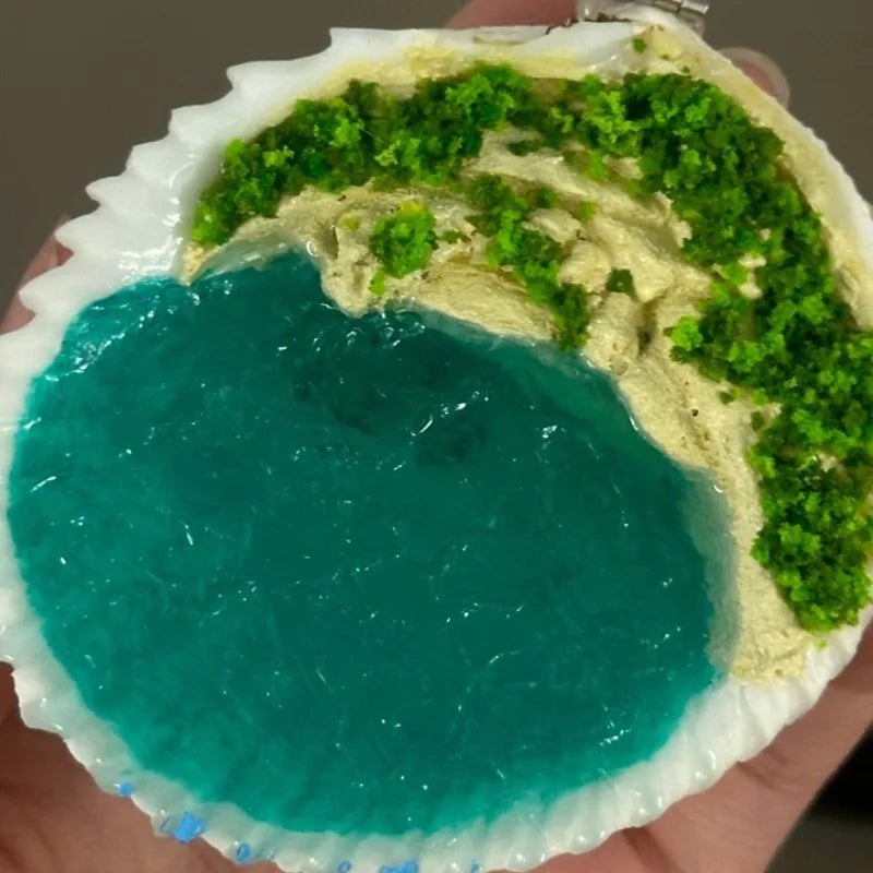 Portable Luxury Ring Box – Aesthetic Wedding Ring & Trinket Storage Case A shell-shaped decorative object with a bright turquoise blue lower section, pale beige upper section, and textured green areas along the top, held in a hand against a neutral background.