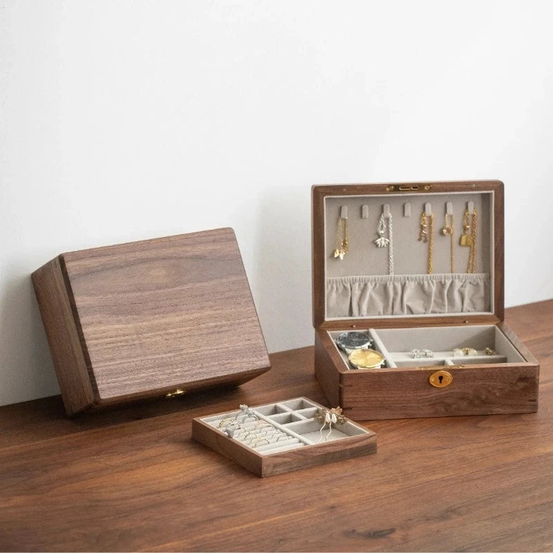 Solid Wood Travel Jewelry Box with Lock – Portable Jewelry Organizer A wooden jewelry organizer set shown on a wooden tabletop against a white wall. One closed box stands on the left, one open box on the right displays hanging hooks, compartments, and a small lock, and a smaller tray sits in front.