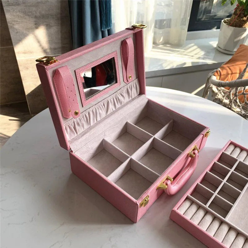Leather Travel Jewelry Box with Mirror – Double-Layer Jewelry Organizer A pink faux-leather jewelry organizer case shown open on a white table, with a matching tray placed beside it. The case has a mirror on the inside lid, multiple interior compartments, ring rolls, and gold-tone hardware, photographed in a bright indoor setting.