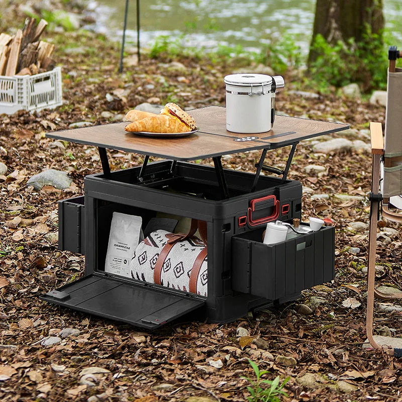 Folding Storage Box – Wooden Lid Camping & Trunk Organizer A black outdoor storage table and storage box set set up in a campsite environment on a leaf-covered ground near water. The table has a wooden top with a white canister and a pastry on it, and the open storage box below contains gear and a patterned blanket.