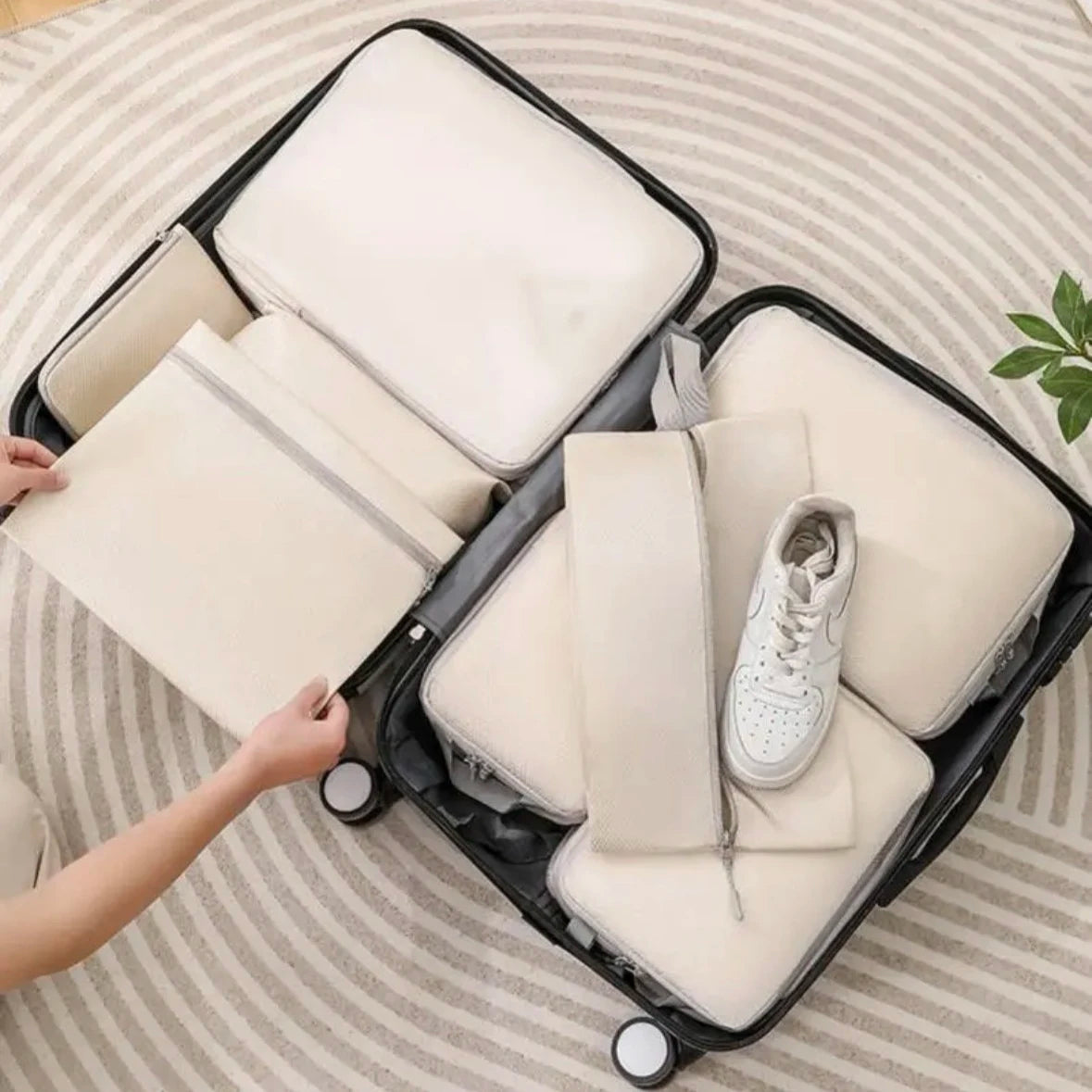 Compression Packing Cubes Set – Space-Saving Luggage Organizers A black hard-shell suitcase opened on a striped rug, filled with multiple cream-colored packing cubes and a white sneaker. A person's hands are visible on the left, adjusting one of the packing cubes.
