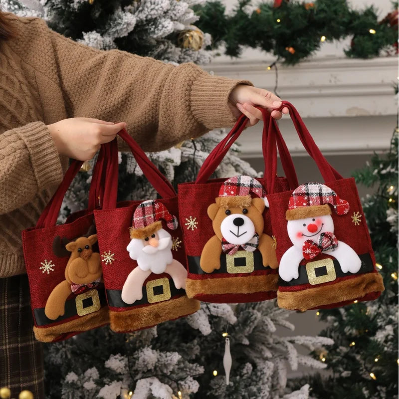 8-Pack Christmas Linen Gift Bags – Santa Candy & Treat Bags Four red Christmas-themed tote bags with plush animal faces are held by a person wearing a brown knit sweater in front of a decorated Christmas tree. The bags feature teddy bears, a Santa Claus figure, a bear with a plaid hat, and a snowman with a plaid hat.