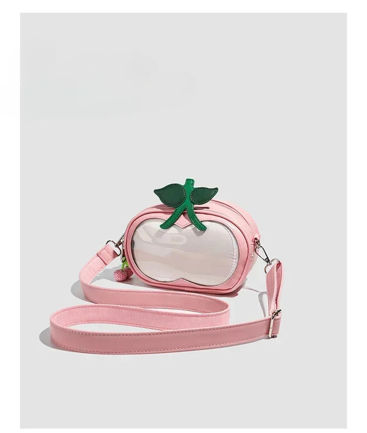 Apple-Shaped Crossbody Phone Purse – Lightweight Shoulder Bag A pink faux-leather crossbody bag designed to look like a peach, featuring a clear front window, a green leaf accent on top, and a long matching pink shoulder strap, shown against a light gray background.
