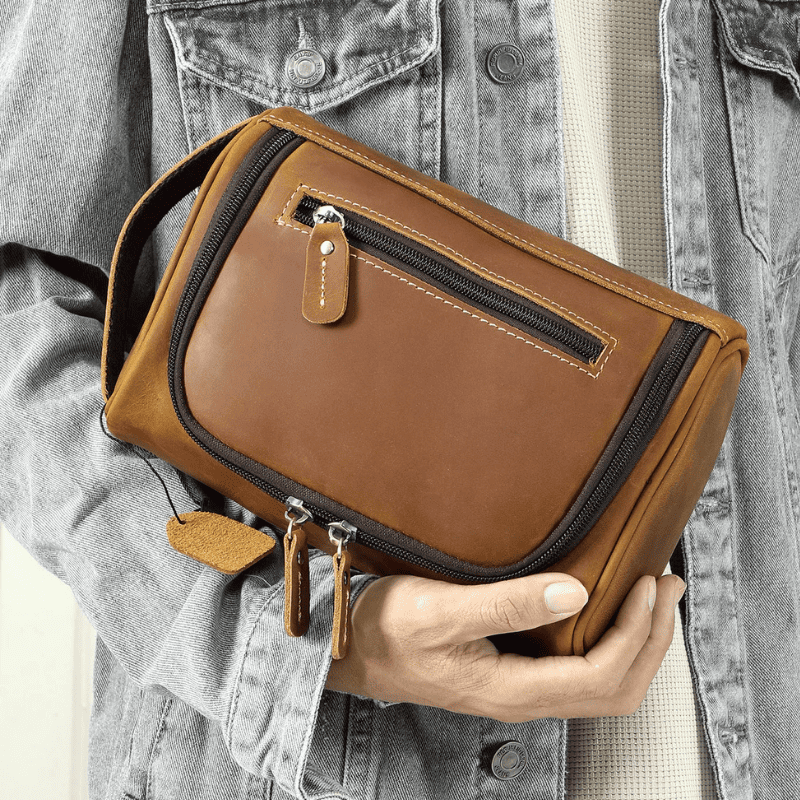 Top-Grain Leather Toiletry Bag for Men – Vintage Travel Organizer Pouch A person in a gray denim jacket holds a brown leather toiletry bag with black zippers and tan leather pull tabs.