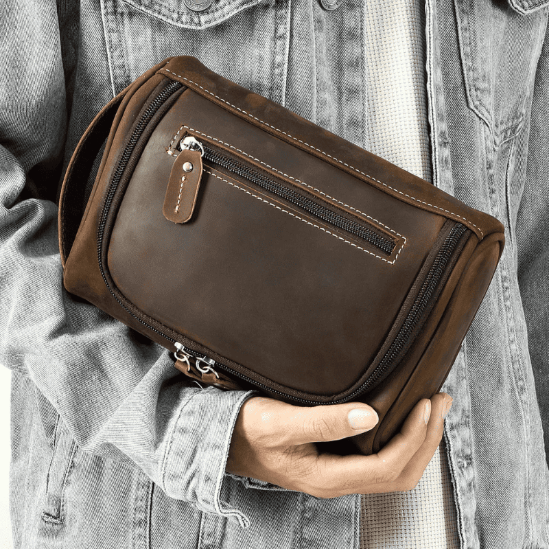 Top-Grain Leather Toiletry Bag for Men – Vintage Travel Organizer Pouch A person in a light gray denim jacket holds a dark brown leather zippered pouch bag. The bag features a top zipper with a leather pull tab and contrast stitching.