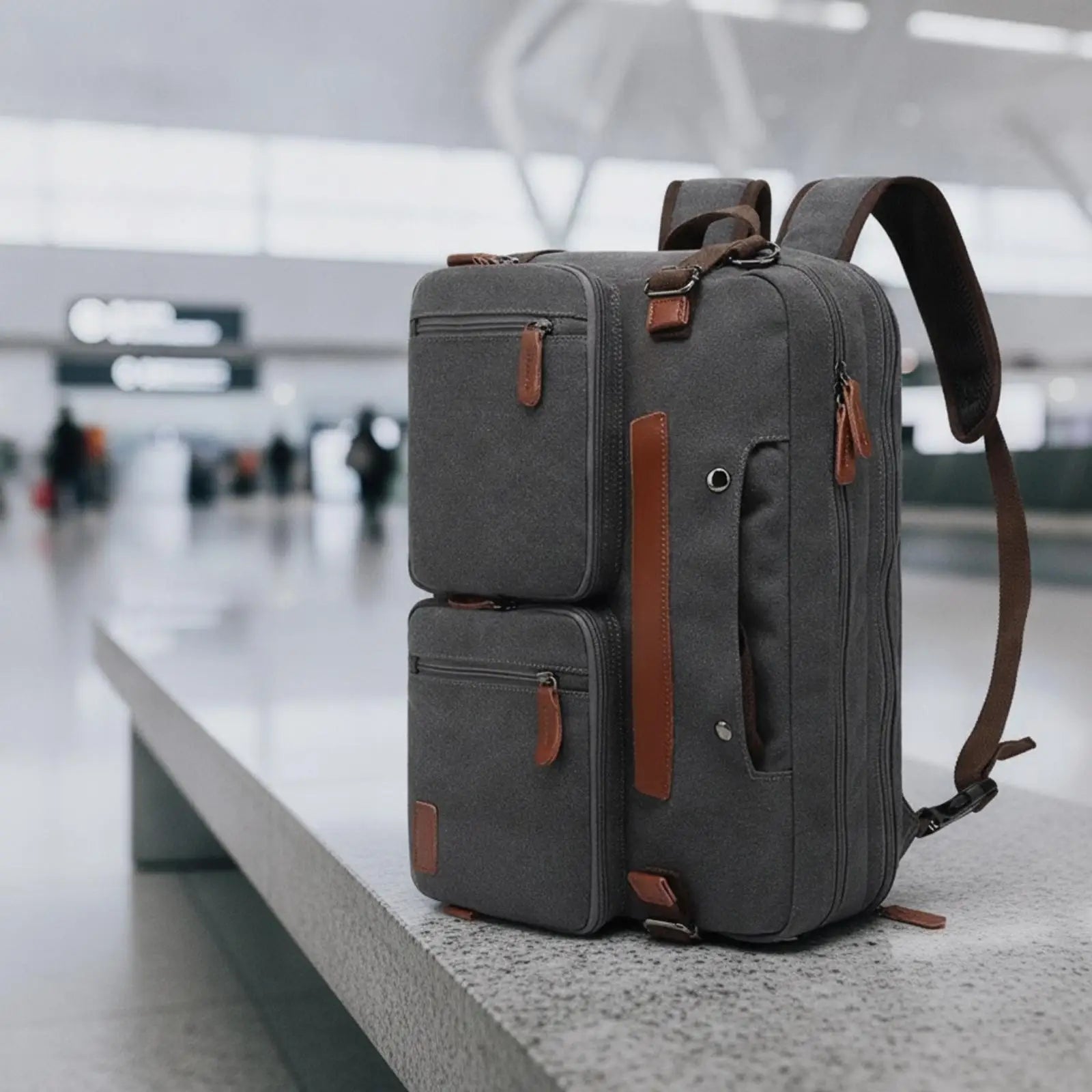 Convertible Laptop Briefcase Backpack – Business Office & Travel Bag A dark gray canvas backpack with brown leather accents sits on a concrete ledge in a blurred airport terminal.