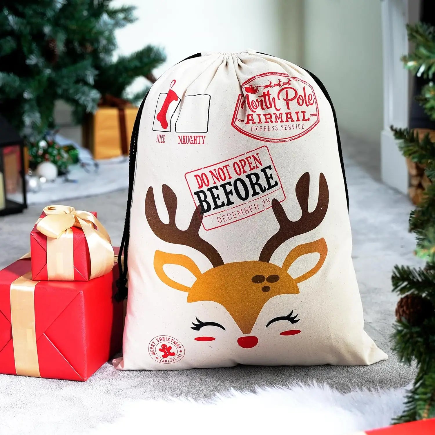 Reusable Christmas Santa Gift Sacks – 3-Pack Canvas Bags A cream-colored Christmas gift sack with a cartoon reindeer face and red 'North Pole Airmail' stamps, styled next to wrapped presents on a holiday floor with a blurred Christmas tree in the background.