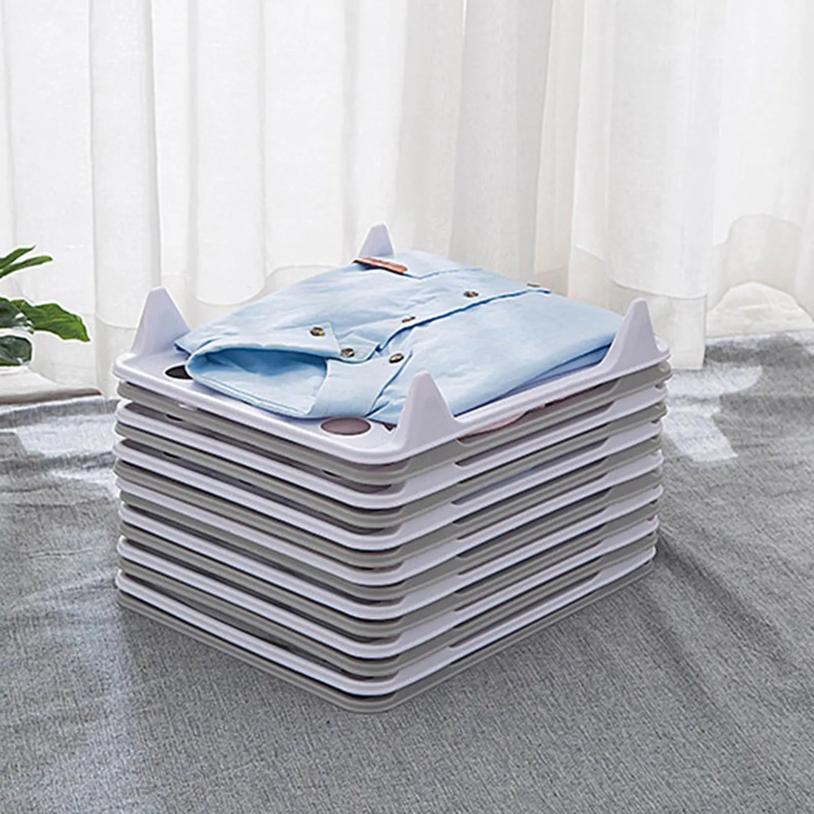 Stackable Clothes Organizers – 5-Piece Interlocking Shirt & T-Shirt Storage A stack of white plastic under-bed storage drawers with triangular legs, shown holding folded light blue shirts. The drawers are arranged on a gray textured blanket in front of white sheer curtains, with a small green plant visible on the left.