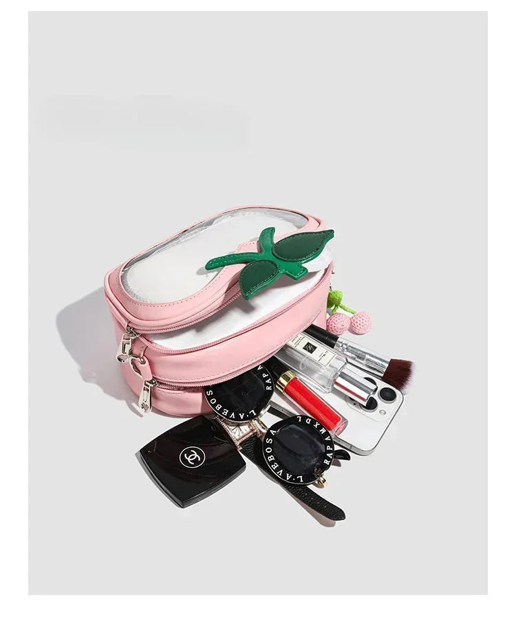 Apple-Shaped Crossbody Phone Purse – Lightweight Shoulder Bag A pink faux-leather cosmetic bag with a clear zippered top and a green strawberry patch, shown open with various makeup items like lipstick, brushes, and a compact mirror spilling out on a light gray background.