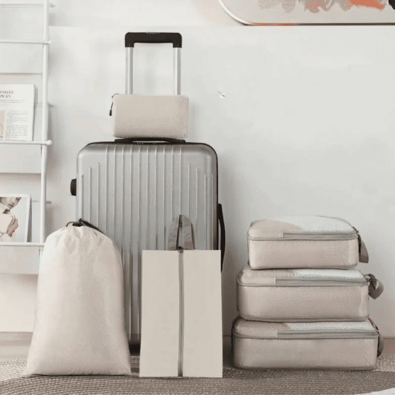 Compression Packing Cubes Set – Space-Saving Luggage Organizers A travel luggage set arranged against a light wall, including a silver hardshell suitcase with the telescoping handle extended, three beige fabric storage bags, and two zippered packing cubes stacked on the right.