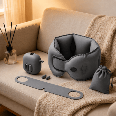 A gray inflatable neck pillow set arranged on a beige sofa with a cream throw blanket, including the main inflatable neck support, a matching gray case, a long gray strap, small earplugs, and a small drawstring pouch. A reed diffuser is visible in the background.