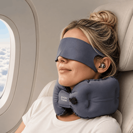 A woman resting on an airplane seat with a gray sleep mask and matching neck pillow around her eyes and neck. The neck pillow has a visible 'EVERYDAY PULL' logo.