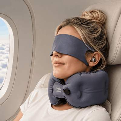 A woman resting on an airplane seat with a gray sleep mask and matching neck pillow around her eyes and neck. The neck pillow has a visible 'EVERYDAY PULL' logo.