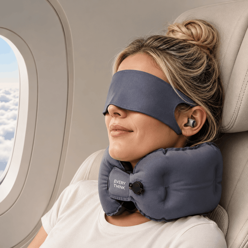 A woman resting on an airplane seat with a gray sleep mask and matching neck pillow around her eyes and neck. The neck pillow has a visible 'EVERYDAY PULL' logo.