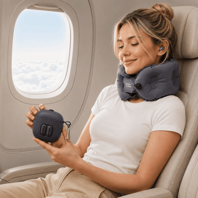 A woman sitting in an airplane seat uses a dark gray neck pillow and earbuds, while holding a matching dark gray portable charging case near the window.