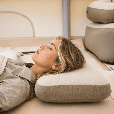A woman lies on her side with her eyes closed, resting her head on a textured light beige pillow. The scene is set in a warm, minimalist indoor environment with neutral-toned cushions in the background.