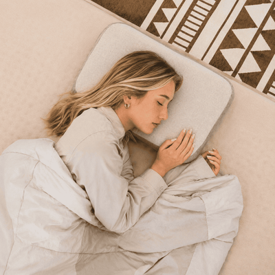 A woman sleeping soundly on a light gray pillow, wrapped in a beige blanket on a bed with a patterned rug.