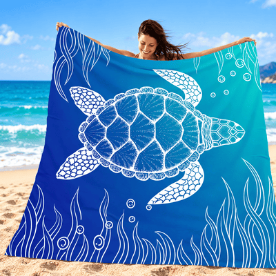 A woman on a sandy beach holds up a large square blue towel or blanket featuring a white sea turtle and underwater wave design. The background shows the ocean, sky, and beach.