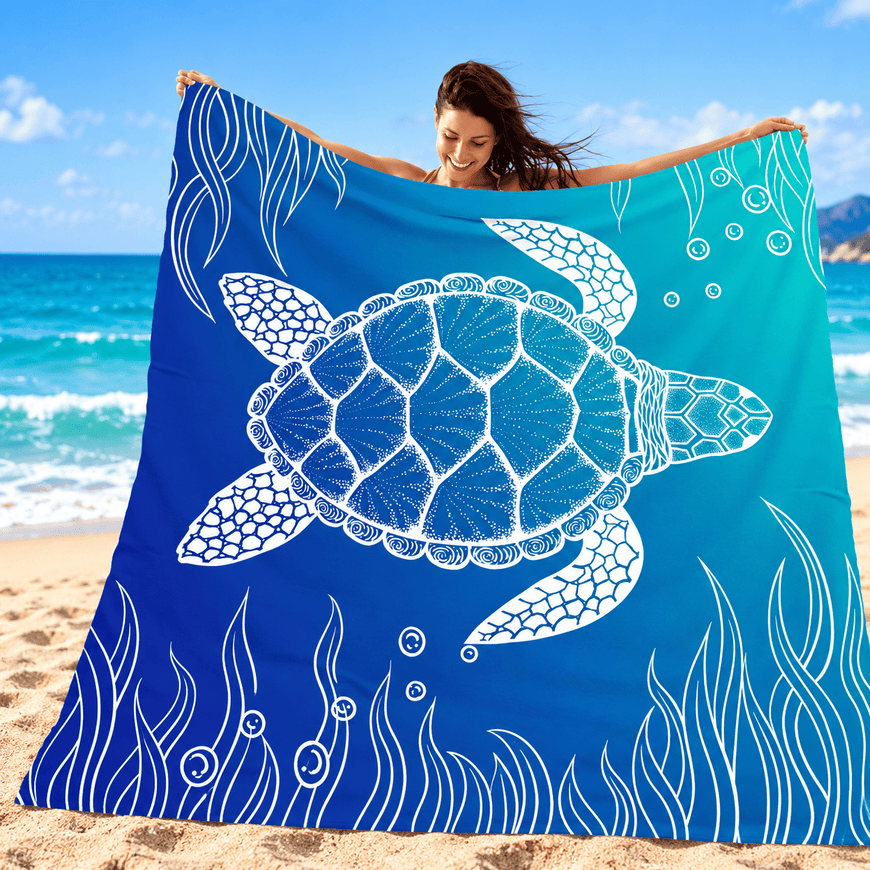 A woman on a sandy beach holds up a large square blue towel or blanket featuring a white sea turtle and underwater wave design. The background shows the ocean, sky, and beach.