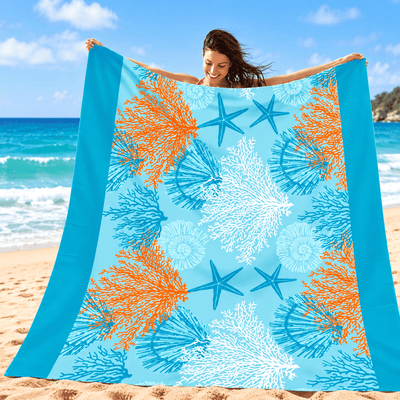 A woman stands on a sandy beach holding up a large square beach towel or blanket. The fabric has a bright turquoise blue border and a central print featuring orange, blue, and white starfish and coral motifs on a light blue background.