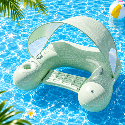 A pale mint green inflatable pool float shaped like a whale or dolphin, with a curved canopy overhead, a built-in footrest, and clear side windows, floating in bright blue swimming pool water with palm leaves and a beach ball visible.
