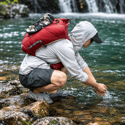 A person crouching in a rocky stream near a waterfall, wearing a light gray hooded jacket, dark shorts, gray slip-on shoes, and a large red backpack.