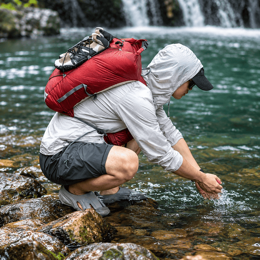 A person crouching in a rocky stream near a waterfall, wearing a light gray hooded jacket, dark shorts, gray slip-on shoes, and a large red backpack.