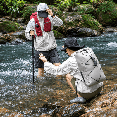 Two hikers cross a rocky stream in a forest. One standing hiker wears a light gray waterproof jacket and a red backpack, while the other crouching hiker in the foreground wears a white waterproof jacket and a large light gray backpack with black straps.