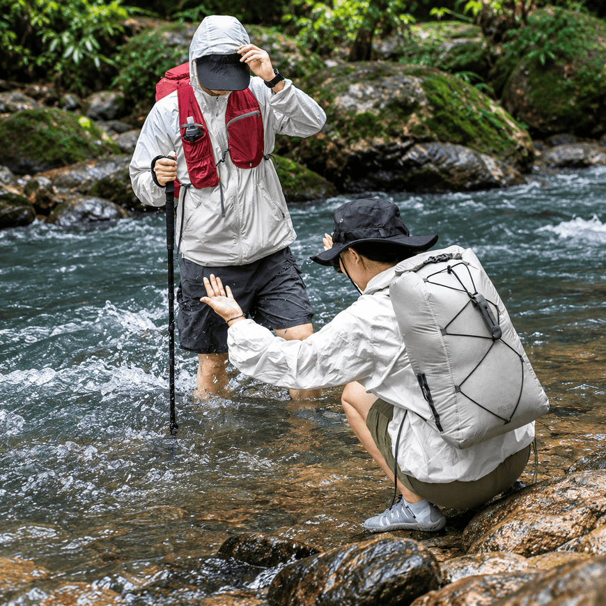 Two hikers cross a rocky stream in a forest. One standing hiker wears a light gray waterproof jacket and a red backpack, while the other crouching hiker in the foreground wears a white waterproof jacket and a large light gray backpack with black straps.