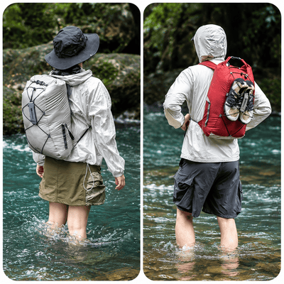 Two people standing in a river, shown from behind, wearing waterproof backpacks. The left person wears a light gray waterproof jacket and shorts, while the right person wears a white waterproof jacket and dark gray shorts.