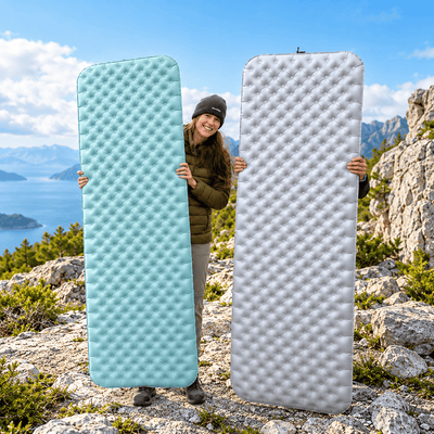 Two people standing outdoors on a rocky mountain with a lake and sky behind them, each holding a large inflatable camping sleeping pad. One pad is light aqua blue and the other is light gray, shown side by side for color comparison.