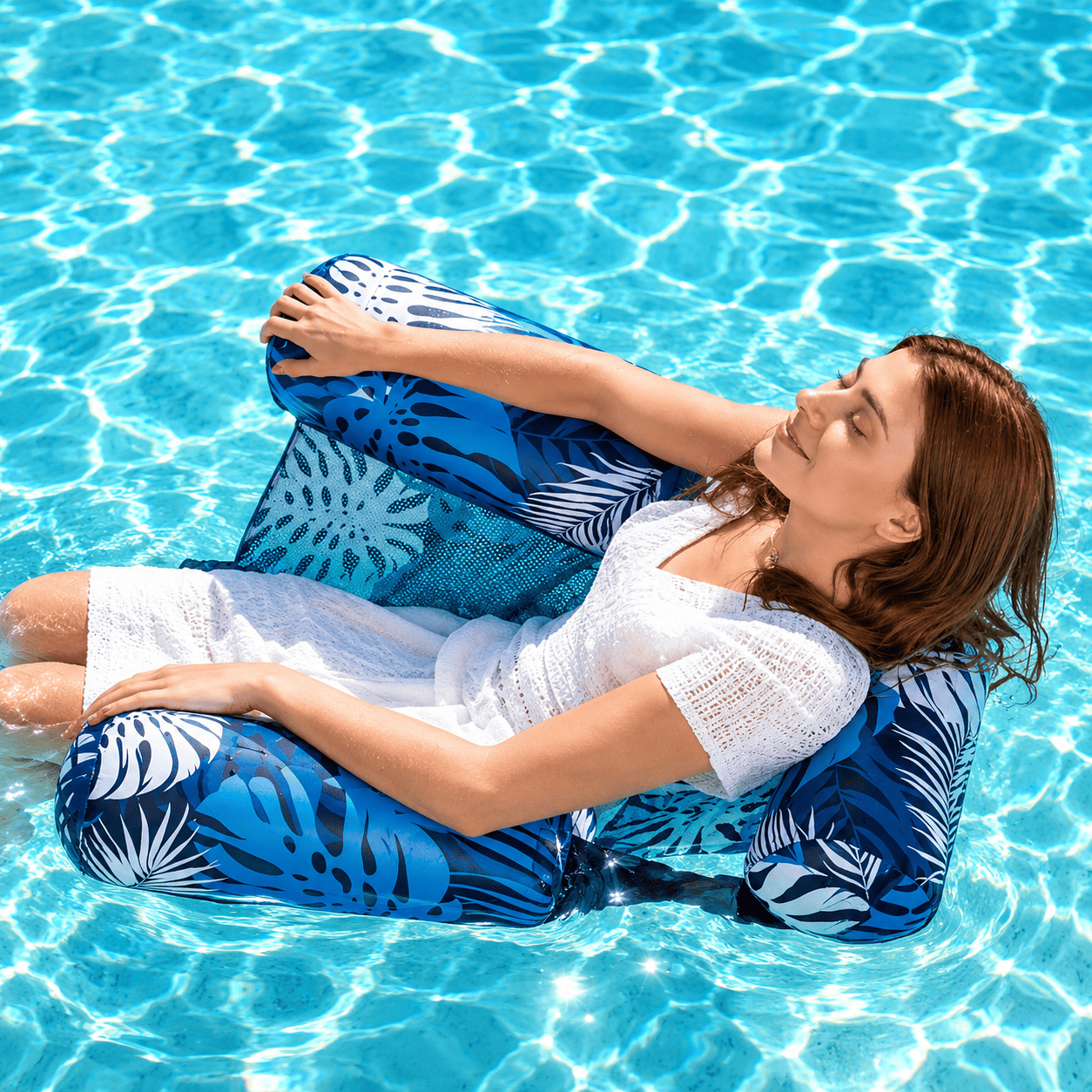 A woman reclines on a blue and white palm-patterned pool float in a bright swimming pool.