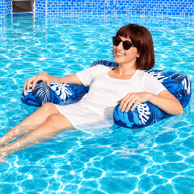A woman wearing sunglasses and a white dress relaxes in a swimming pool on a blue and white tropical-patterned inflatable pool float.