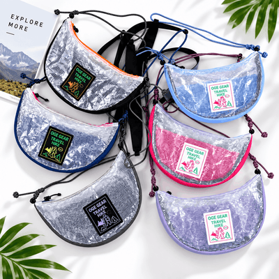 Six small crescent-shaped pouches with a shiny sequin-like silver surface are arranged on a white background. Each pouch has a different trim color: orange, black, blue, purple, pink, and lavender, and a front patch reads 'OCE GEAR TRAVEL ADS'.