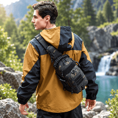 A man wearing a yellow and black outdoor jacket stands in a mountainous, forested landscape with a waterfall and turquoise water in the background, carrying a black tactical sling backpack across his chest.