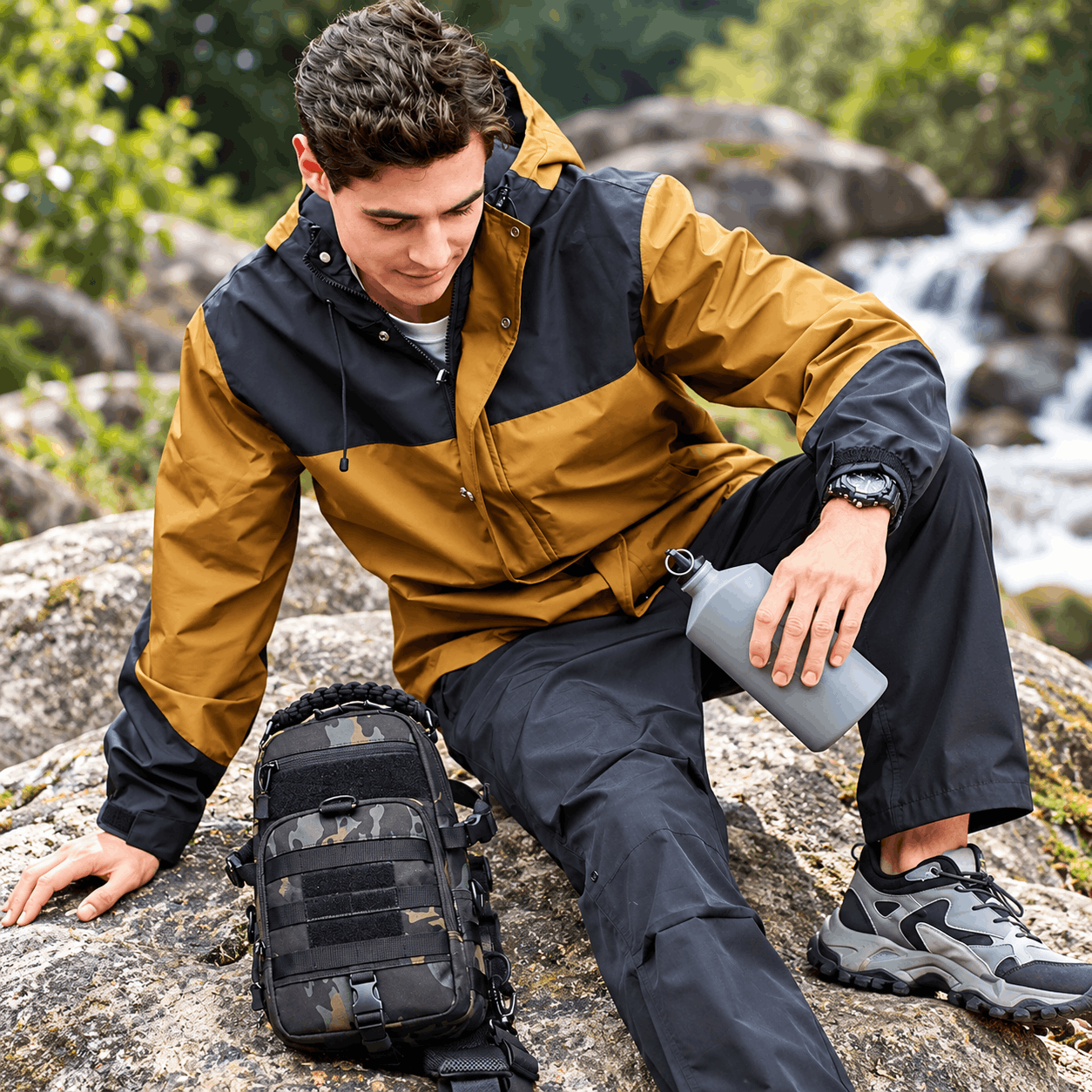 A man sitting on a large rock outdoors near a flowing stream, wearing a two-tone tan and navy hooded jacket, dark pants, and hiking shoes. A black tactical-style shoulder bag with a camouflage pattern rests on the rock beside him, and he holds a gray water bottle.