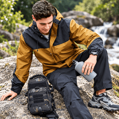 A man sitting on a large rock outdoors near a flowing stream, wearing a two-tone tan and navy hooded jacket, dark pants, and hiking shoes. A black tactical-style shoulder bag with a camouflage pattern rests on the rock beside him, and he holds a gray water bottle.
