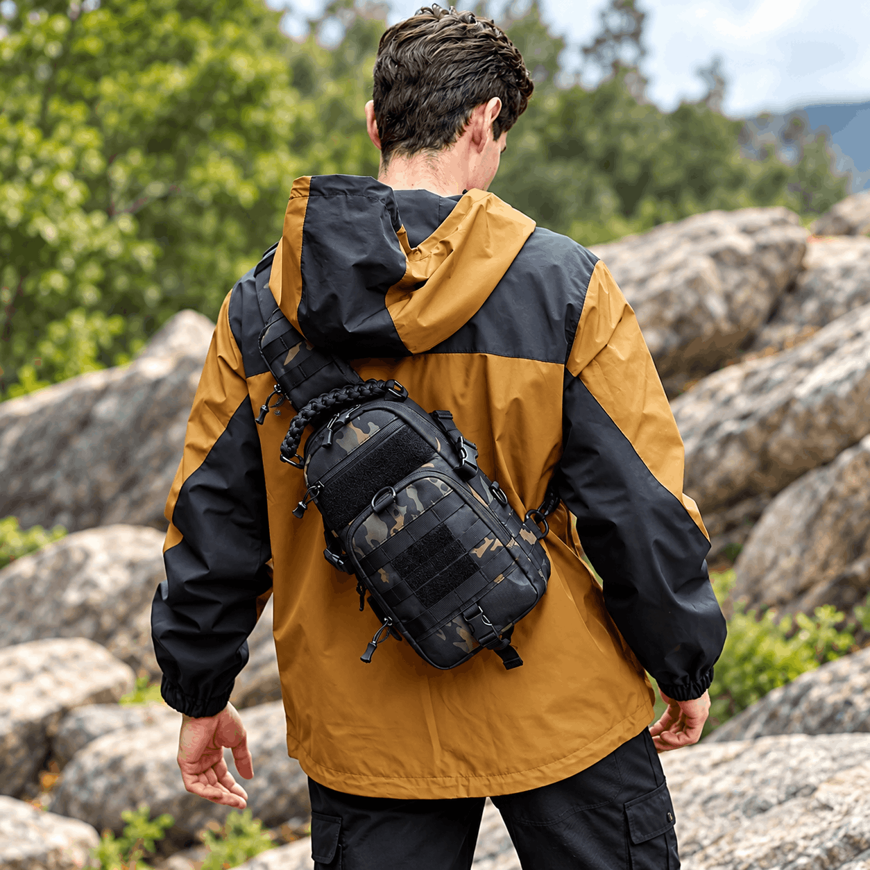 A man viewed from behind wearing a black and mustard yellow hooded jacket, carrying a black tactical sling backpack with a camouflage front pocket, outdoors among large rocks and green trees.