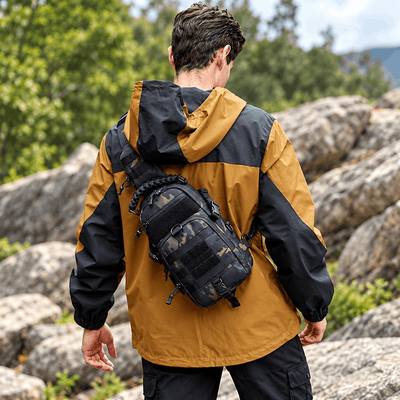 A man viewed from behind wearing a black and mustard yellow hooded jacket, carrying a black tactical sling backpack with a camouflage front pocket, outdoors among large rocks and green trees.