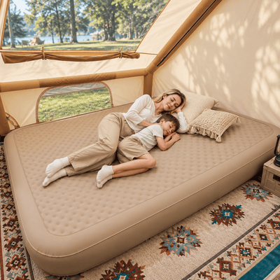 A woman and a child relax on a large beige camping air mattress inside a tent, with a patterned rug underneath and a scenic outdoor view through the tent flap.
