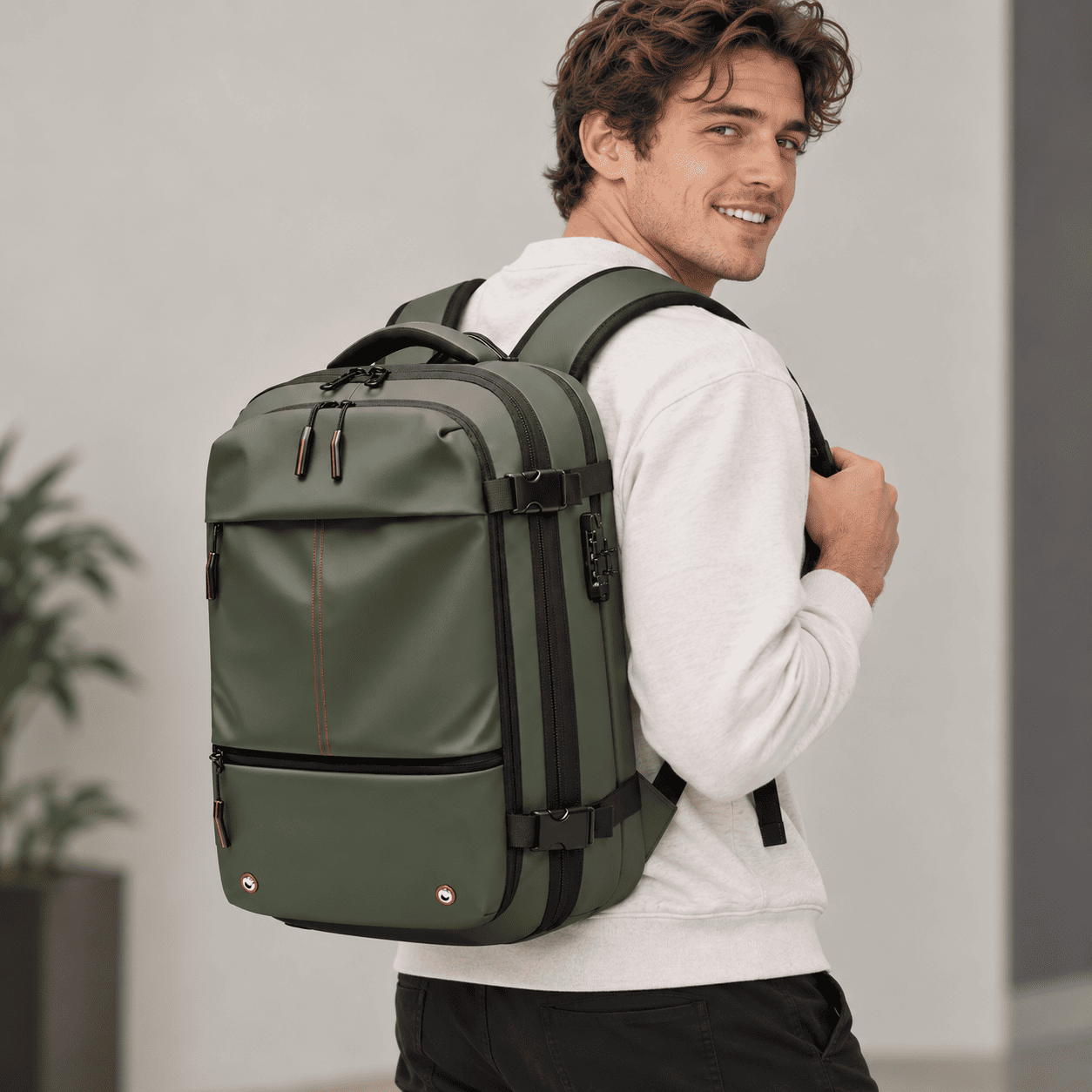 A man wearing a white sweatshirt and black pants carries an olive green backpack with black straps and trim, photographed in a clean indoor setting.