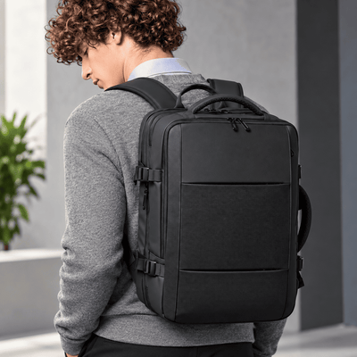 A man wearing a gray sweater and light blue collared shirt is shown from behind, wearing a black multi-compartment backpack. The setting is a modern outdoor corridor with gray walls and a small green plant on the left.