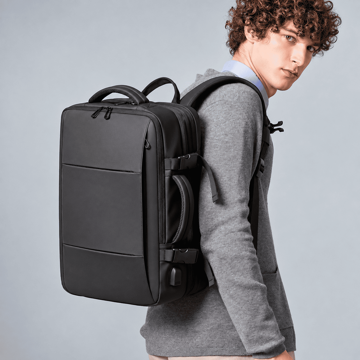 A man wearing a gray hoodie and light blue shirt carries a black laptop backpack over his shoulder, photographed against a solid light gray studio background.