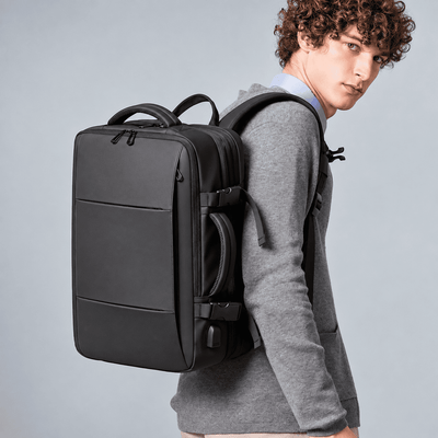 A man wearing a gray hoodie and light blue shirt carries a black laptop backpack over his shoulder, photographed against a solid light gray studio background.