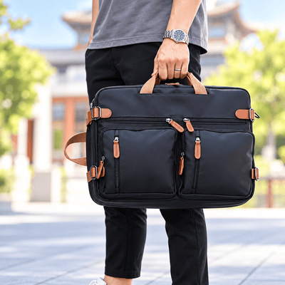 A man in a gray t-shirt and black pants holds a black nylon laptop briefcase with tan leather handles and zipper pulls in an outdoor setting with a blurred traditional building in the background.