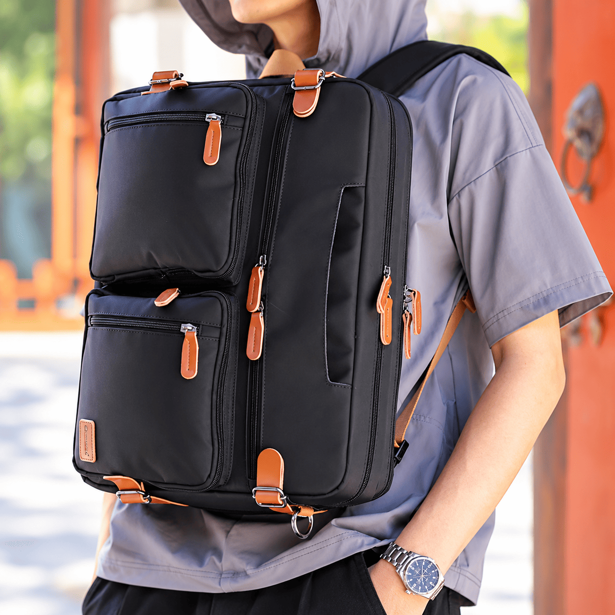 A person wearing a gray short-sleeve shirt carries a black multi-compartment backpack with tan leather-like zipper pulls and straps in an outdoor setting with warm-toned architectural elements.